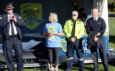 Man in a police uniform holding a microphone while a woman in a sports apparel and two more man watch over