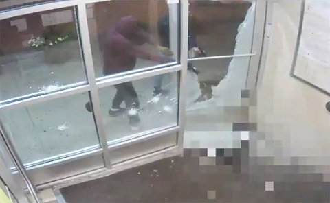 Two people in hoods holding handguns behind of shattered glass door