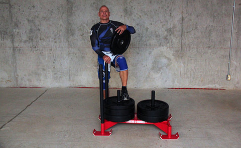 Man dressed in a blue sports outfit holding a cylindrical weight, with one foot on a metal sled that's loaded with more weights