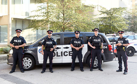 Group of men and women, wearing police uniforms, standing in a parking lot in-front of various police vehicles.